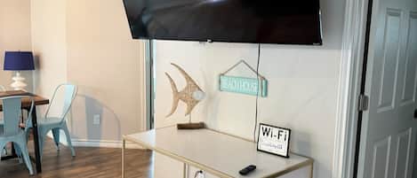 A modern living room with a flat-screen TV mounted on the wall, a small table with a sign indicating Wi-Fi availability, a colorful rug, and a door leading to another room.
