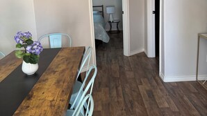 A dining area with a wooden table, white chairs, and a vase of flowers. A hallway leads to a bedroom with a bed and a nightstand.
