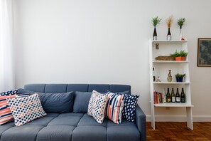 A modern living room with a grey sofa, patterned cushions, a white shelving unit with decorative items, and a wooden floor.