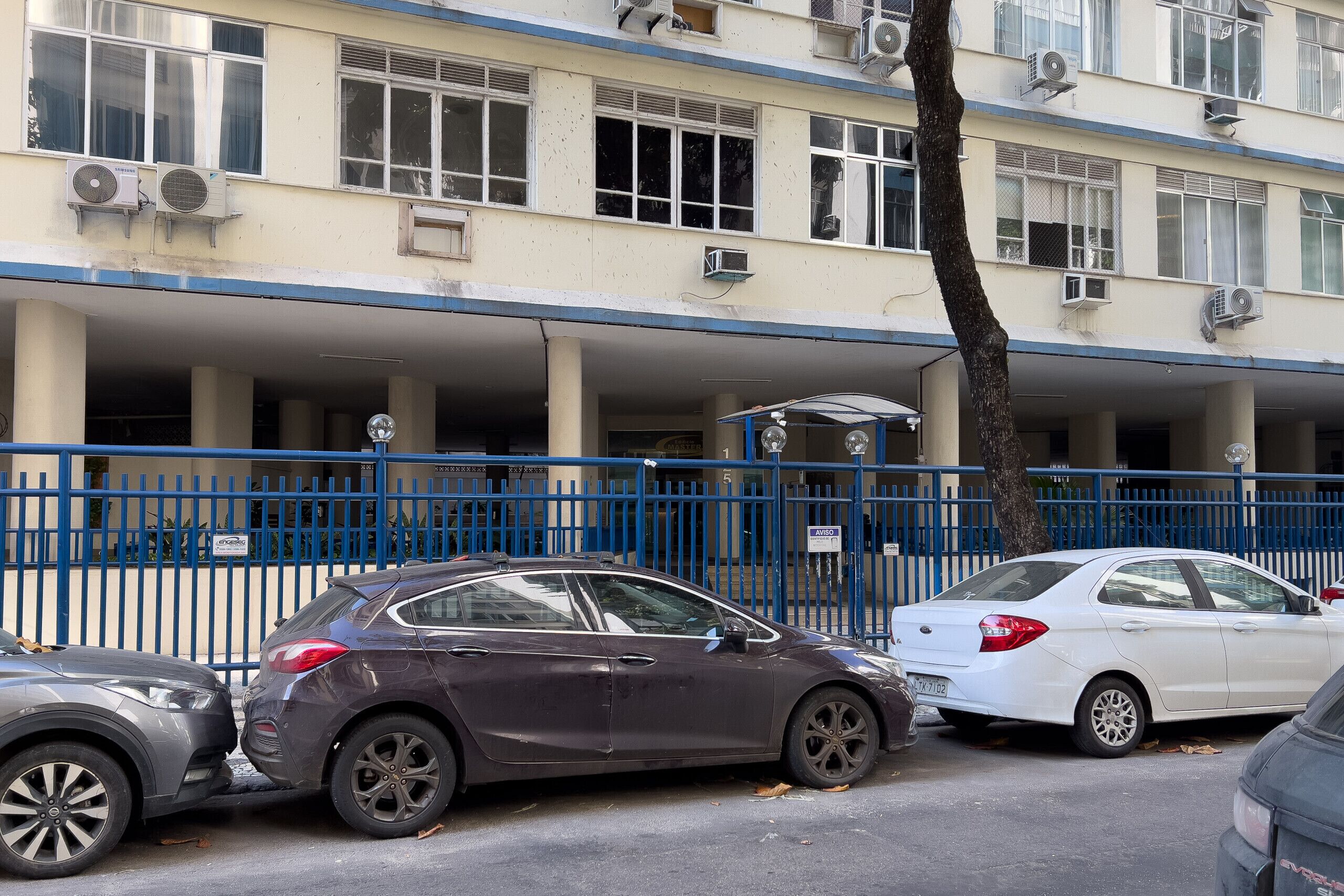 A building with multiple air conditioning units, a blue metal fence, and parked cars.