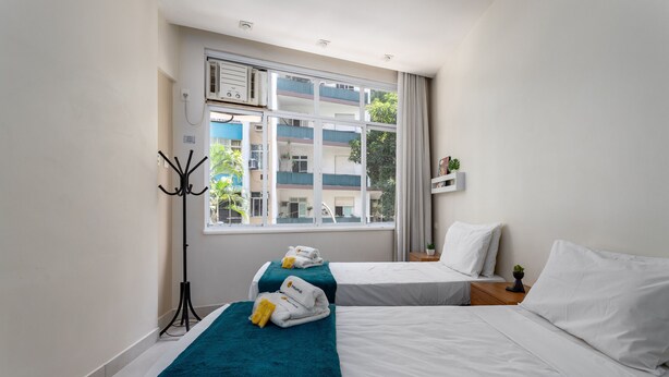 A hotel room with two beds, a window with a view of buildings, a coat rack, and a small plant on the nightstand.