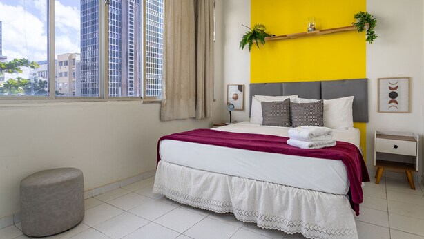 Ein modernes Schlafzimmer mit großem Fenster, einem Bett mit weißer und roter Bettdecke, einem grauen Kopfteil und einem kleinen Nachttisch mit Pflanze.