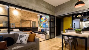 Una sala moderna con un sofá gris, una mesa de comedor con sillas, y una cocina con un armario amarillo y electrodomésticos de acero inoxidable.