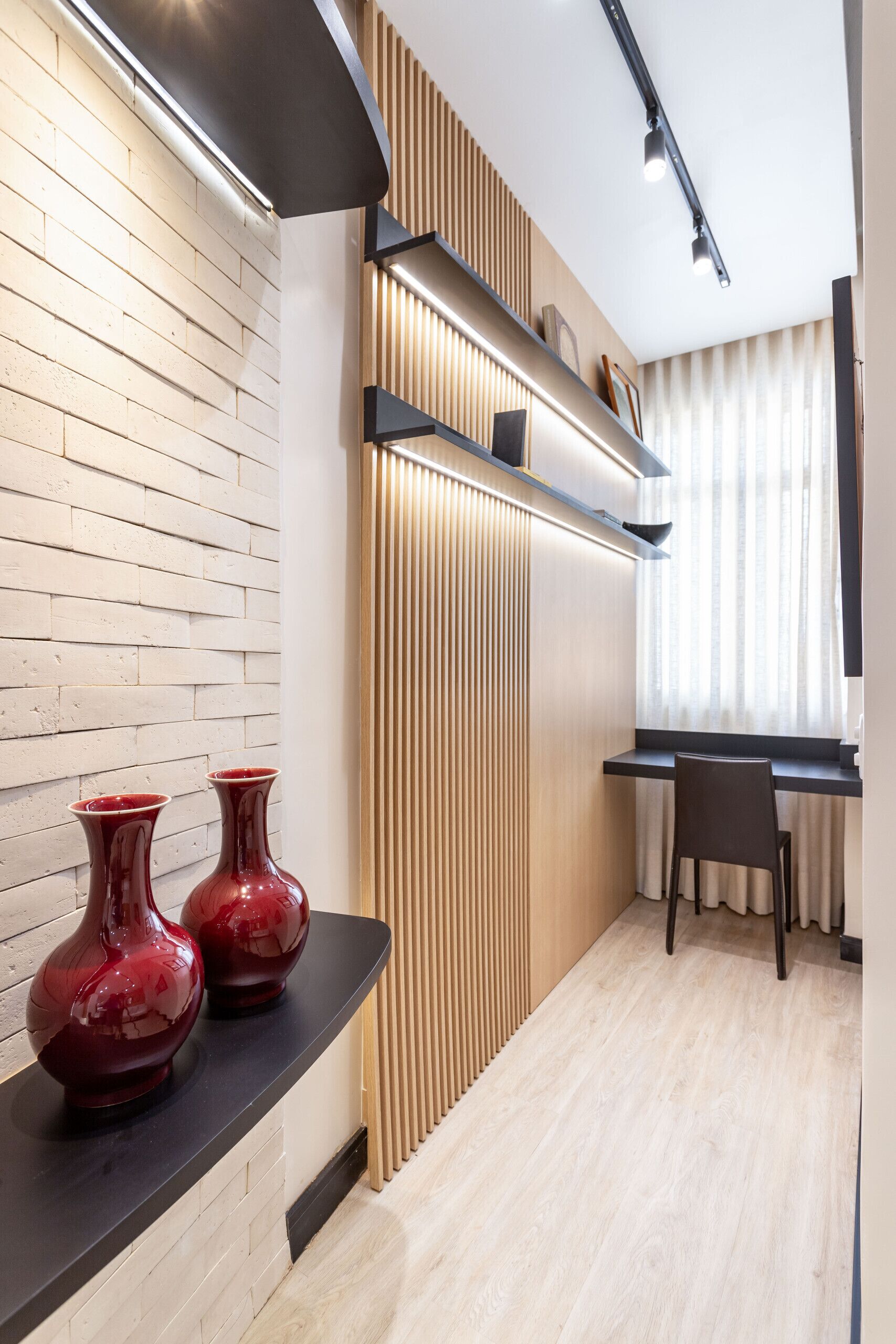 A modern interior with a wooden wall, a black countertop with two red vases, a wooden slatted panel, a black chair, and a desk with a window.