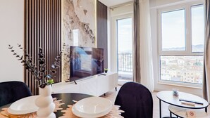 Un salon moderne comprenant un téléviseur, une table à manger et des chaises, une petite plante, et offrant une vue sur la ville grâce à de grandes fenêtres.