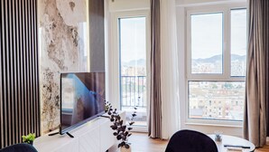 Un salon moderne doté d’un téléviseur à écran plat, d’une table à manger et offrant une vue sur la ville grâce à de grandes fenêtres.