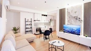 Un salon moderne avec un canapé, une table à manger et des chaises, un téléviseur fixé au mur et une cuisine en arrière-plan.
