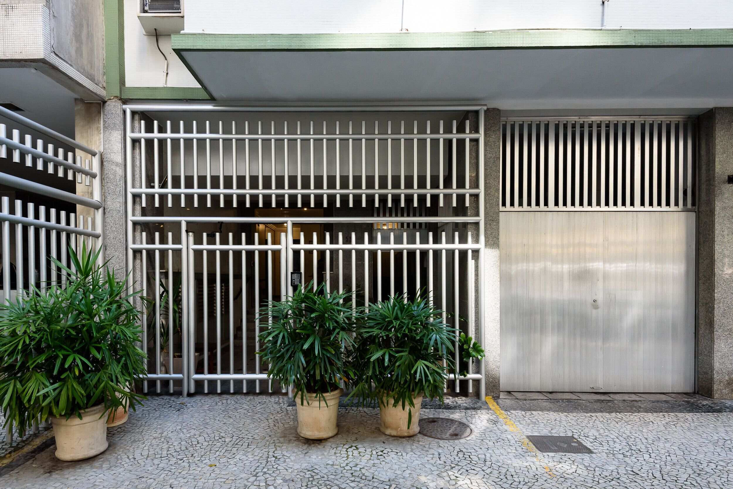 Uma entrada gradeada com barras de metal, plantas em vasos e uma porta de metal fechada.