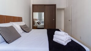Ein ordentlich bezogenes Bett mit weißen Leinen, einer schwarzen Tagesdecke und einem Stapel weißer Handtücher. Ein Holz-Kopfteil und gemusterte Kissen sind zu sehen. Im Hintergrund ist ein Holzschrank erkennbar.