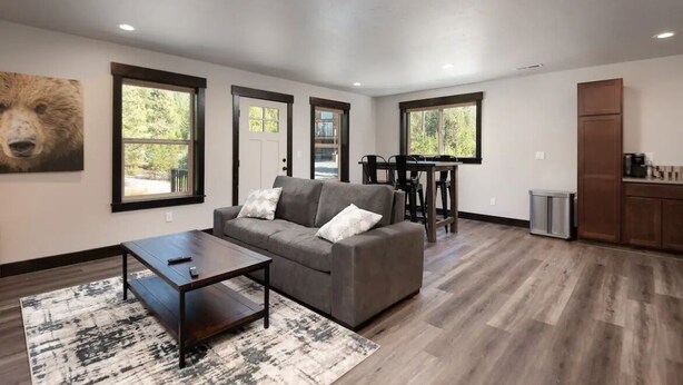 Un salon moderne avec un canapé gris, une table basse et un tapis. De grandes fenêtres offrent une vue sur la verdure environnante.