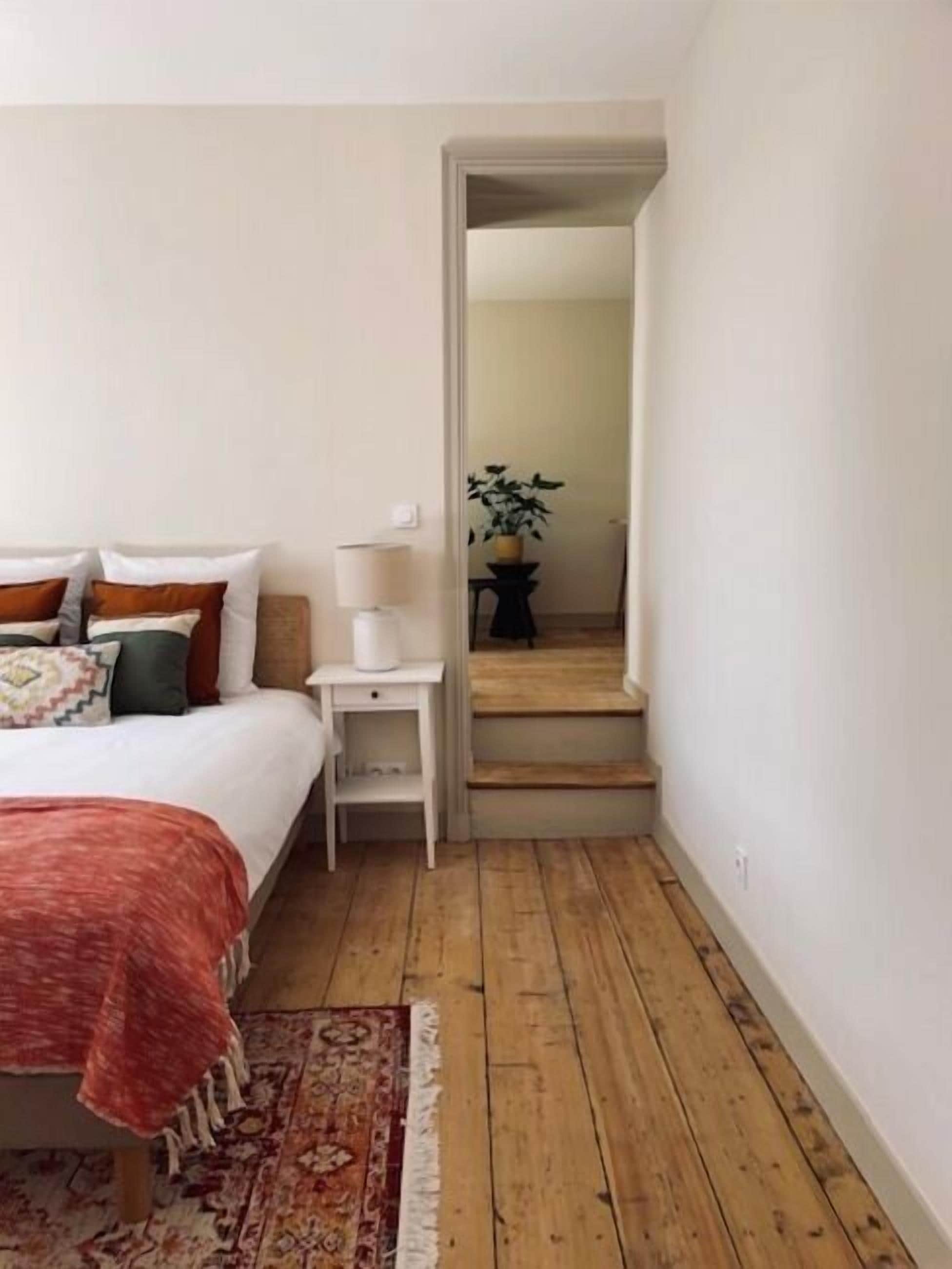 Un dormitori amb un llit, una tauleta de nit, una làmpada, una planta i una escala que porta a una altra habitació.