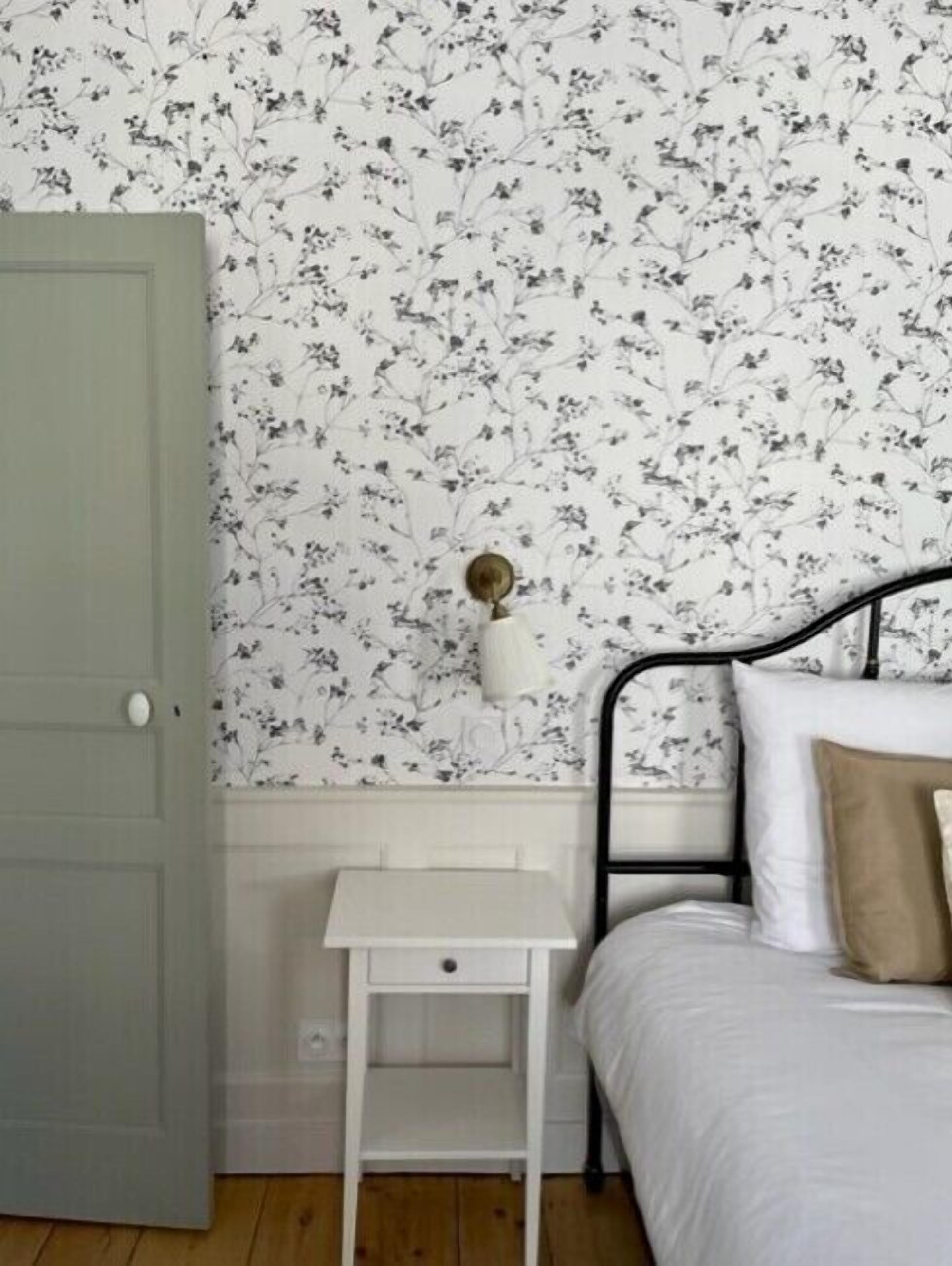 Une chambre avec un papier peint à motifs floraux, une table de chevet blanche, un lit avec des coussins blancs et beiges, et une porte verte.