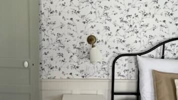 Une chambre avec un papier peint à motifs floraux, une table de chevet blanche, un lit avec des coussins blancs et beiges, et une porte verte.