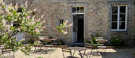 Une cour bordée de murs en pierre, avec une porte en bois, et des tables et des chaises disposées pour créer un espace de détente en plein air.