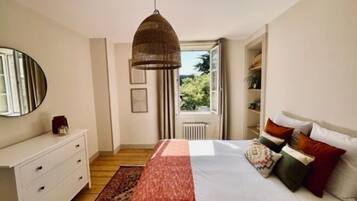 A bedroom with a bed, a dresser, a mirror, a window with a view of greenery, and a hanging lamp.
