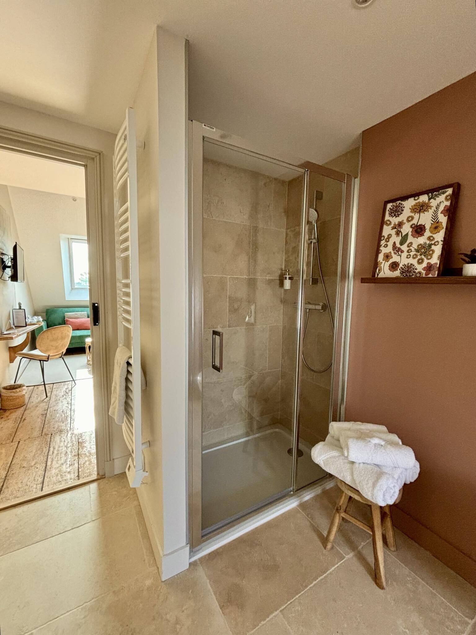 A modern bathroom with a glass-enclosed shower, a wooden stool with a towel, and a framed floral artwork on the wall.