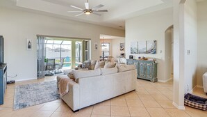 A spacious living room with a large sofa, a ceiling fan, and a view through glass doors to the outside. - Cozy Cape Coral Home: 8 Mi to Marina! (Cape Coral)