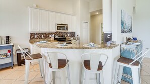 A kitchen with white cabinets, a granite countertop, and a stainless steel microwave. There are four bar stools with wooden seats and white metal frames. - Cozy Cape Coral Home: 8 Mi to Marina! (Cape Coral)
