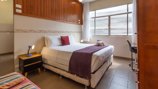 Una habitación de hotel con cama, mesita de noche, lámpara y ventana con persianas.