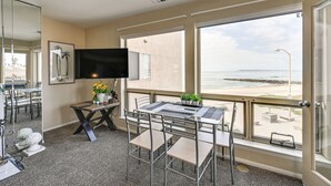 A room with a large window overlooking the beach, a flat-screen TV mounted on the wall, and a table with chairs.