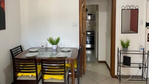 Comedor con una mesa de madera dispuesta para cuatro, sillas con cojines amarillos, una planta pequeña sobre la mesa y una cocina visible a través de una puerta abierta.
