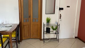Una habitación con puerta de madera, pared blanca, un estante con plantas, un espejo y una mesa con sillas.
