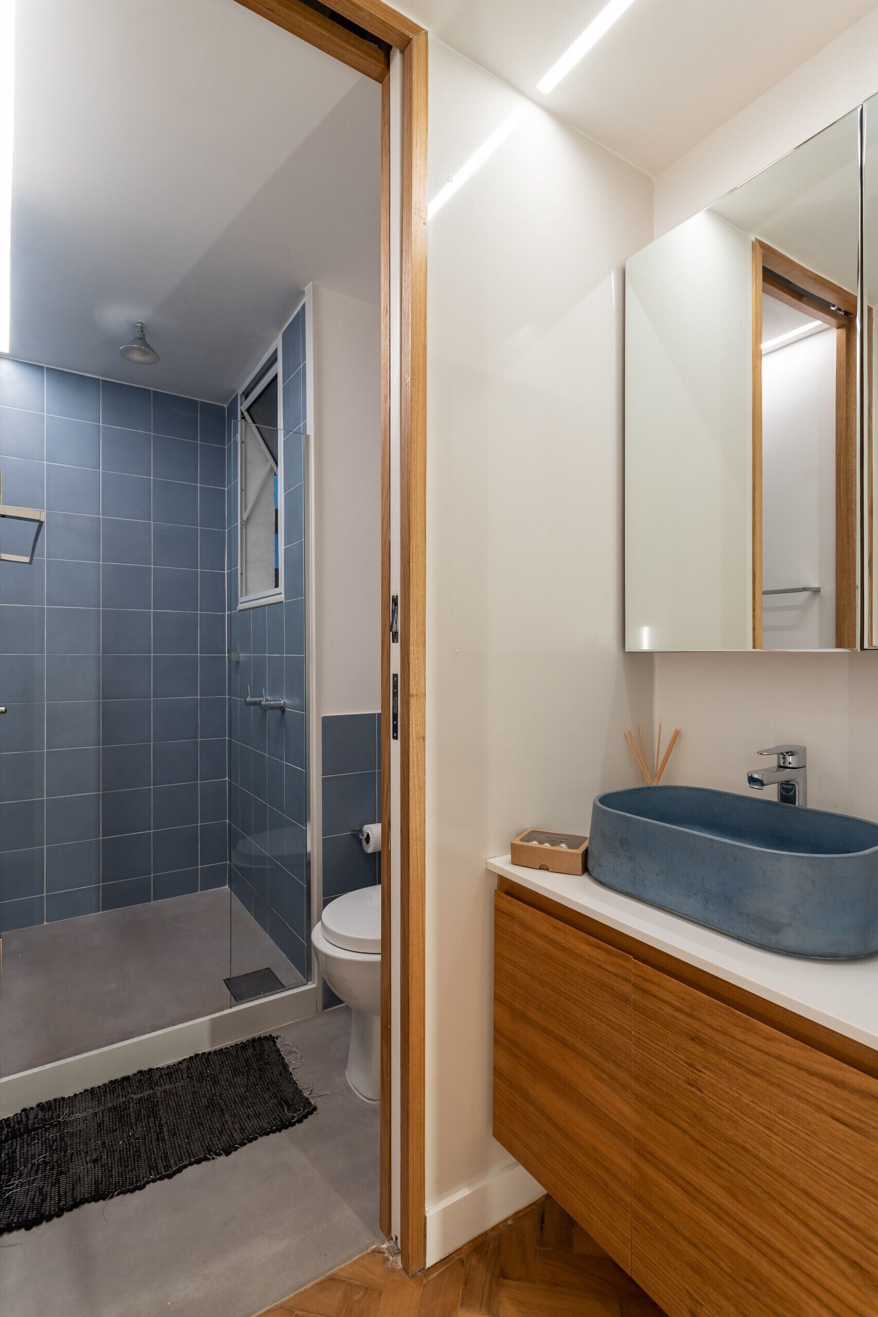 A modern bathroom with a blue-tiled shower, a white toilet, and a wooden vanity with a blue sink.