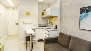 Eine kleine Küche mit weißen Schränken, einer Mikrowelle und einem Spülbecken. Im Wohnbereich befindet sich ein braunes Sofa und ein kleiner Hocker.