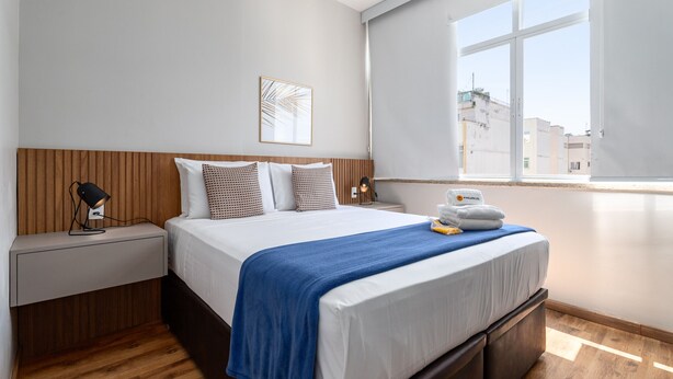 A modern hotel room with a wooden headboard, a large bed with a blue blanket, a bedside table with a lamp, and a window with a city view. - Comfort in Copacabana | 150m from the beach | NSC1203 (Rio de Janeiro)