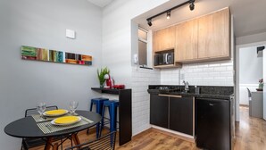 A modern kitchen with black cabinets, a microwave, and a compact refrigerator. There is a dining area with a black table and blue stools. - Comfort in Copacabana | 150m from the beach | NSC1203 (Rio de Janeiro)