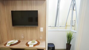 Un comedor con un televisor montado en la pared, una mesa de madera dispuesta para dos personas y una planta en maceta cerca de una ventana.