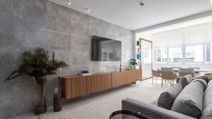 A modern living room with a large flat-screen TV mounted on a grey wall, a wooden reception desk, a grey sofa, and a glass coffee table.