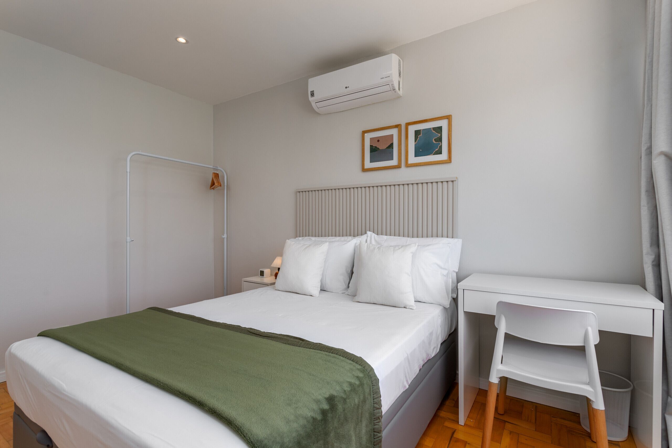 A bedroom with a bed, a desk, a chair, an air conditioner, and two framed pictures on the wall.