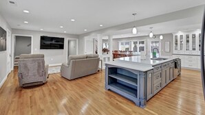 Una sala de estar amplia con un sofá gris y un sillón reclinable, una cocina con una isla grande y armarios blancos, y un comedor con mesa y sillas.