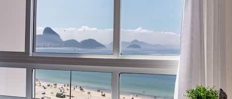 A room with a large window overlooking a beach, a small table, and a chair.