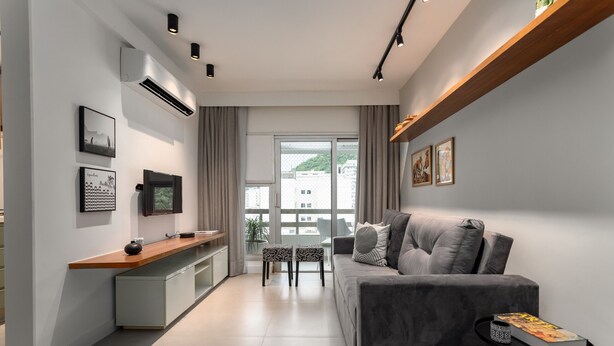 Una sala de estar moderna con un sofá gris, una mesita pequeña y un televisor montado en la pared. Se ve una ventana con cortinas, un ventilador de techo y una pequeña mesita de noche con un libro.