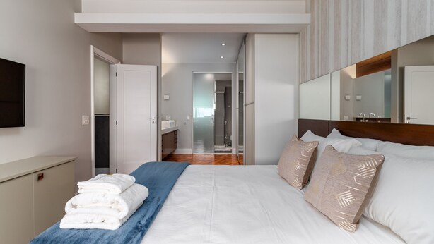 Habitación de hotel moderna con una cama grande, una mesita de noche con toallas, un espejo y vistas al baño y al pasillo.