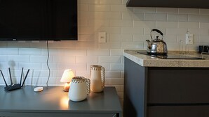 Une cuisine moderne avec un téléviseur à écran plat fixé au-dessus d’un mur en briques blanches, une bouilloire en acier inoxydable posée sur le plan de travail et une petite table agrémentée d’une lampe et d’objets décoratifs.