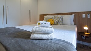 Un lit bien fait, recouvert d’une couverture grise, une pile de serviettes blanches ornées d’un logo représentant un ananas, et une lampe de chevet.