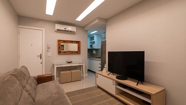 A modern living room with a sofa, a TV on a wooden stand, a small kitchen area, and an air conditioning unit. - 3 minutes from Copacabana beach | STC303 (Rio de Janeiro)