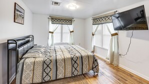 Ein Schlafzimmer mit einem Bett, einem an der Wand montierten Fernseher und einem Fenster mit Vorhängen.