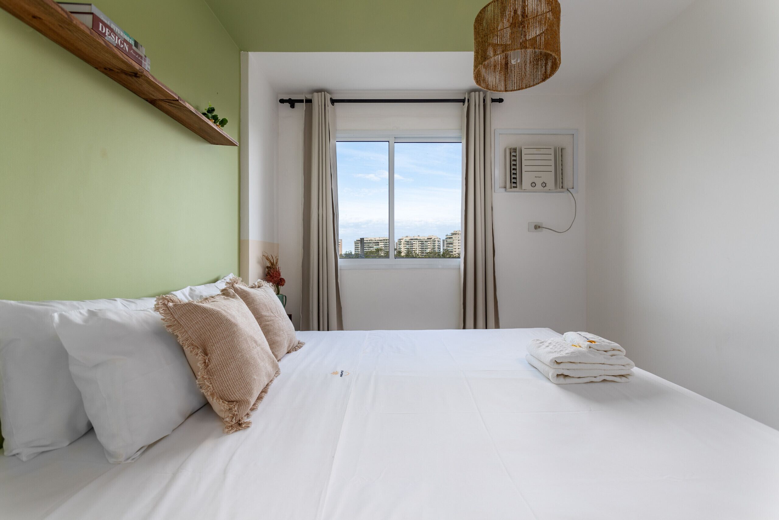 Une chambre à coucher avec un lit, des oreillers, une fenêtre avec des rideaux, un radiateur fixé au mur et une étagère avec des livres.