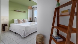 A bedroom with a bed, a bedside table, a lamp, a shelf with books, and a window with a view. - Beauty at Recreio | For 5 peopple | SA201 (Rio de Janeiro)