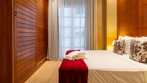 Une chambre d’hôtel avec un mur lambrissé en bois, une grande fenêtre ornée de rideaux légers, un lit avec une literie blanche et des coussins à motifs floraux, et un banc rouge avec des serviettes.