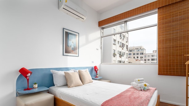 A modern bedroom with a wooden bed, a small bedside table, a red lamp, a window with a view of buildings, and a wall-mounted air conditioner.