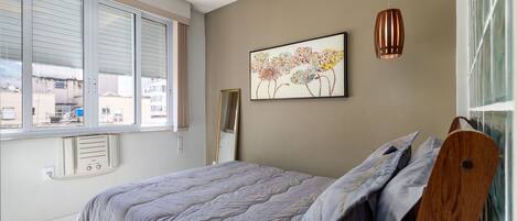 A bedroom with a large bed, a window with blinds, a radiator, and a painting on the wall.