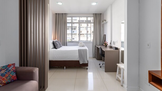 Et moderne hotelværelse med seng, skrivebord, stol og udsigt over byen gennem store vinduer.