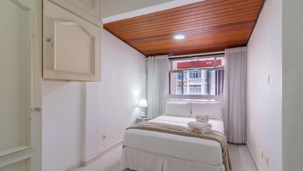 A bedroom with a wooden ceiling, a bed with white bedding, a window with curtains, and a view of a building outside.