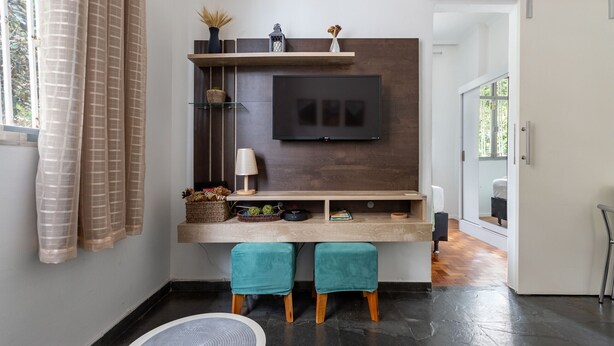 A modern living room with a flat-screen TV mounted on a dark wood panel, teal ottomans, a coffee table, and a small shelf with decorative items.