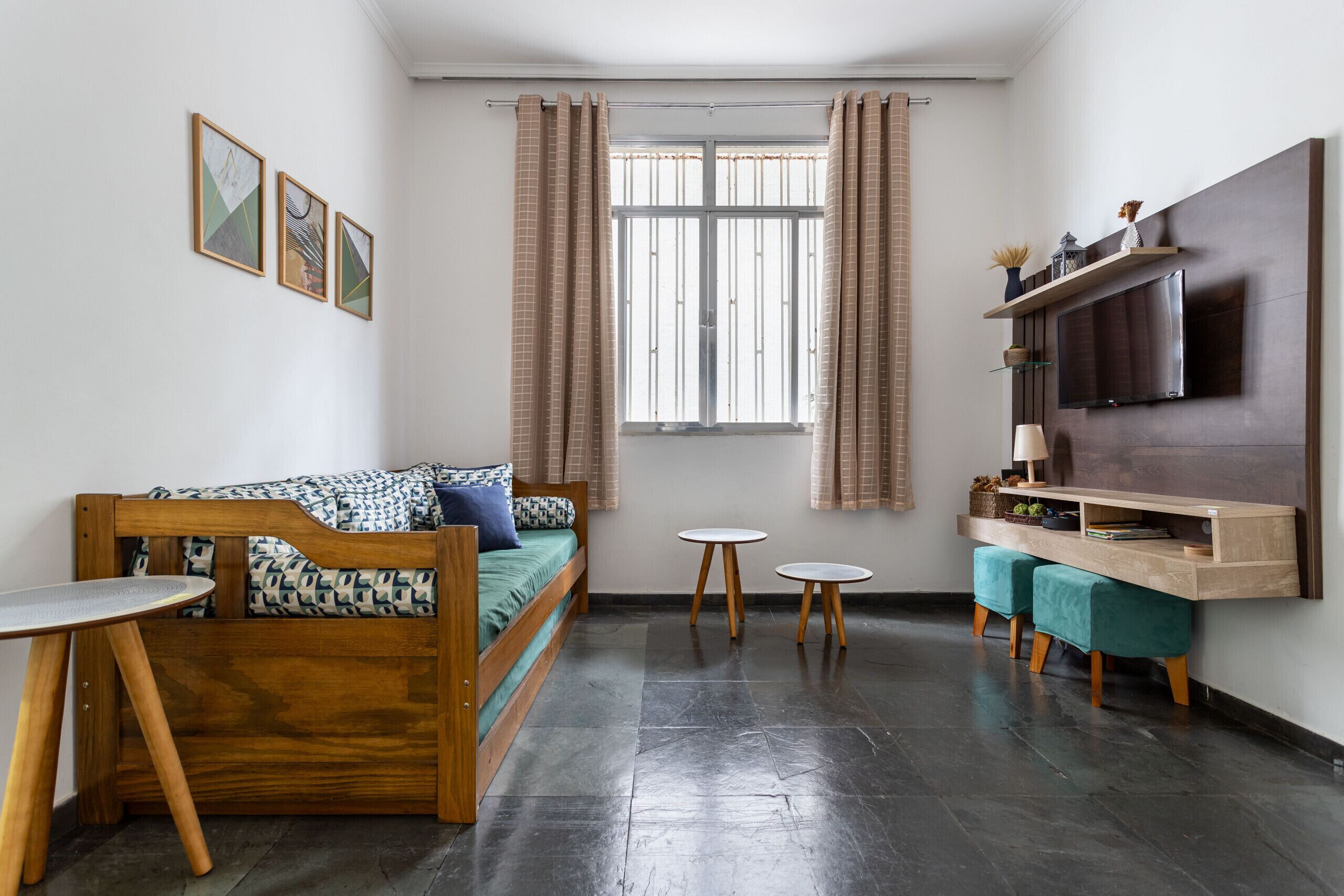 Un espacio habitable compacto con un sofá de madera, una mesita, dos taburetes redondos, un televisor montado en la pared y una ventana con cortinas.